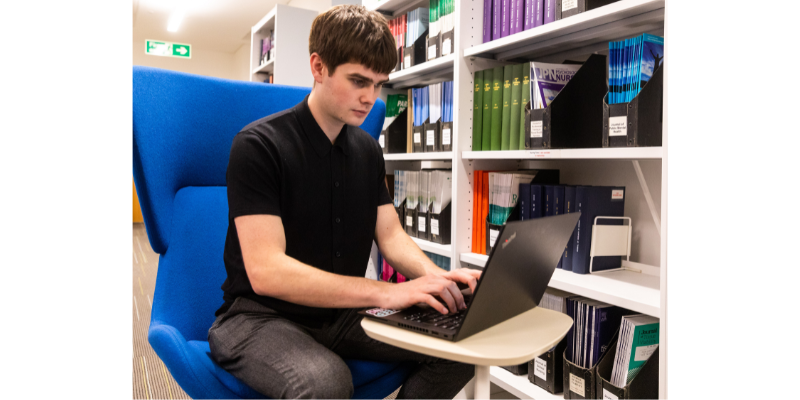 Young man using a laptop beside a shelf of journals in a library