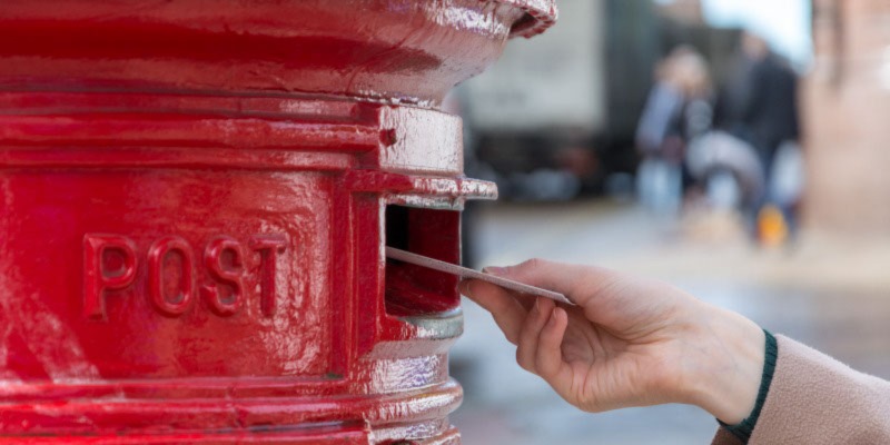 Photo shows white hand posting envelope into red post box