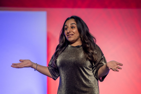 A smiling woman with long dark hair, wearing a shimmering gold top, stands on a stage with a red and purple backdrop, gesturing with her arms wide open.