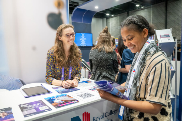 Two women smiling and conversing at an exhibition stand; one stands behind the counter while the other reviews a brochure she is holding.