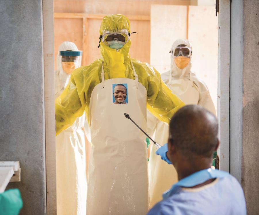 Image of three people wearing full PPE waiting to be sprayed down by a fourth person with their back to us. the person in the front has their portrait without PPE on the front of their apron.
